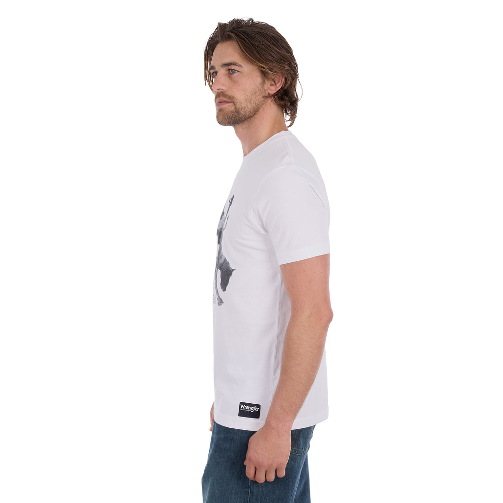A man in profile wearing a white Wrangler Men's Michael Short Sleeve Tee (X6S1557684) with a graphic of a cowboy riding a bucking bronco and the text 'where legendary lives' and a Wrangler logo, paired with blue jeans, against a plain white background.
