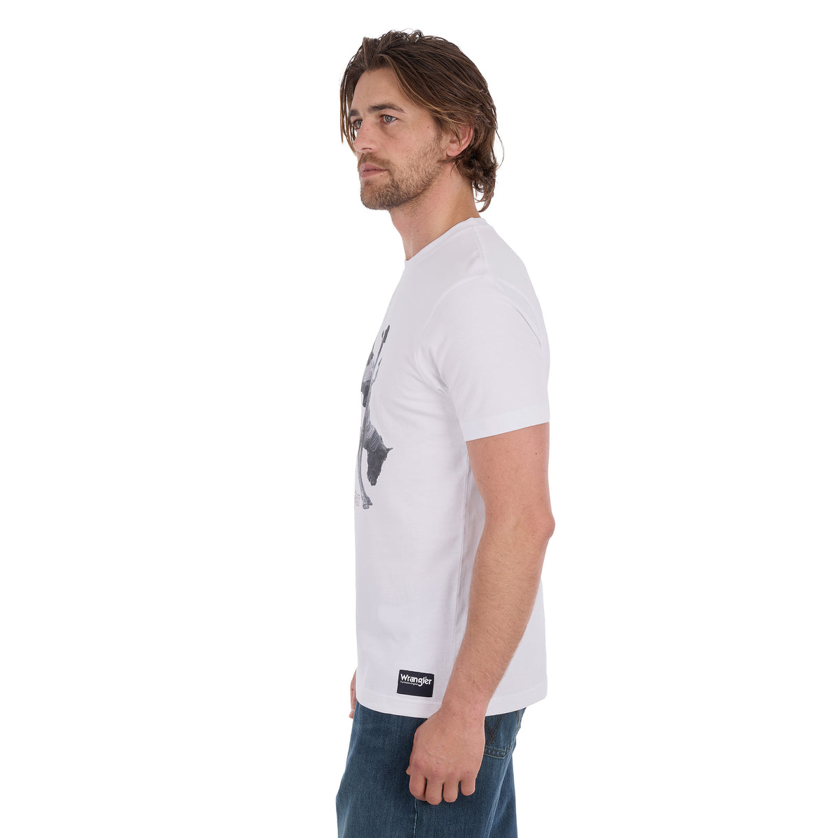 A man in profile wearing a white Wrangler Men's Michael Short Sleeve Tee (X6S1557684) with a graphic of a cowboy riding a bucking bronco and the text 'where legendary lives' and a Wrangler logo, paired with blue jeans, against a plain white background.