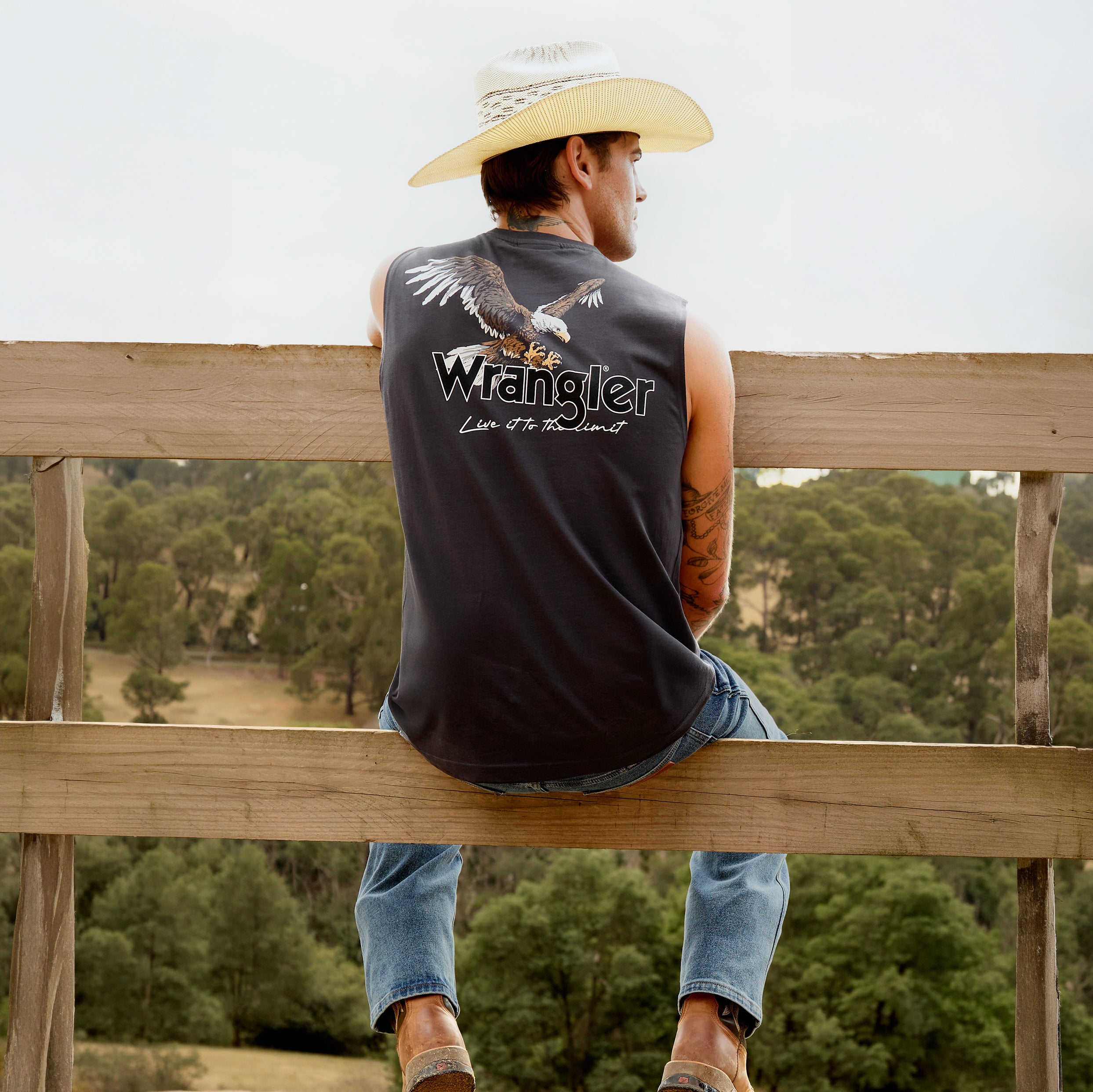 A man with his back to the camera, sitting on a wooden rail fence wearing a Wrangler Men's Paul Muscle Tank with a Wrangler logo and a large print of an eagle on it (XCP1572700) in Charcoal with a pair of denim jeans.