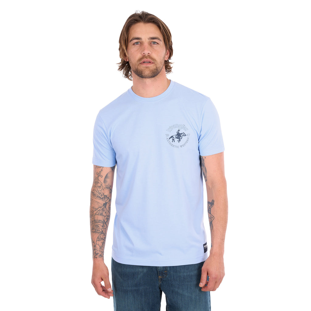 A man wearing a light blue Wrangler Men's Steve Short Sleeve Tee (X6S1557597) with a small circular graphic on the left chest, paired with blue jeans, photographed against a solid white background.