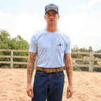 A man wearing a light blue Wrangler Men's Steve Short Sleeve Tee (X6S1557597) with a black cowboy graphic on the left chest, blue jeans, a toffee Wrangler Fletch Belt (XCP1908BLT) and a navy and tan Wrangler Chase High Profile Trucker Cap (XCP1941CAP), standing next to a brown horse in a sandy outdoor arena.