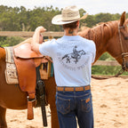 A man wearing a light blue Wrangler Men's Steve Short Sleeve Tee (X6S1557597) with a black cowboy graphic on the back, blue jeans, toffee Wrangler Fletch Belt (XCP1908BLT) Toffee and a Wrangler Hudson Bangora Cowboy Hat (XCP1928HAT), standing next to a brown horse in a sandy outdoor arena.
