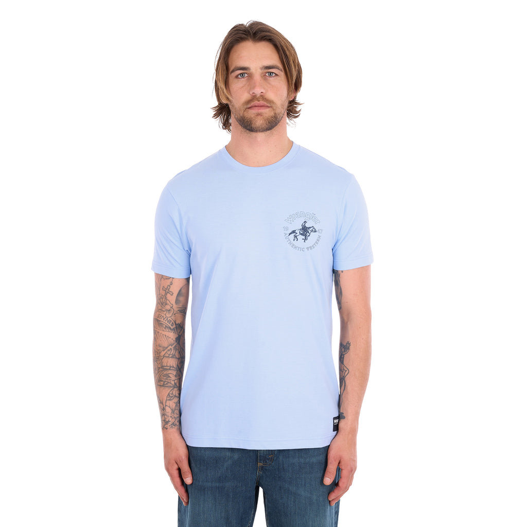 A man wearing a light blue Wrangler Men's Steve Short Sleeve Tee (X6S1557597) with a small circular graphic on the left chest, paired with blue jeans, photographed against a solid white background.