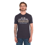 A man wearing a charcoal Wrangler Men's Tim Short Sleeve Tee (X6S1557696) with a Wrangler graphic on the chest, paired with blue jeans, standing against a plain white background.