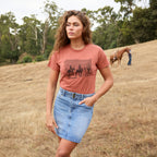 A woman stands in a dry grassy field wearing a copper coloured Wrangler Women's Adele Tee (X6S2598644) with a dark graphic print on the front and a faded blue Wrangler Women's Maisy Denim Skirt (XCP2409723).