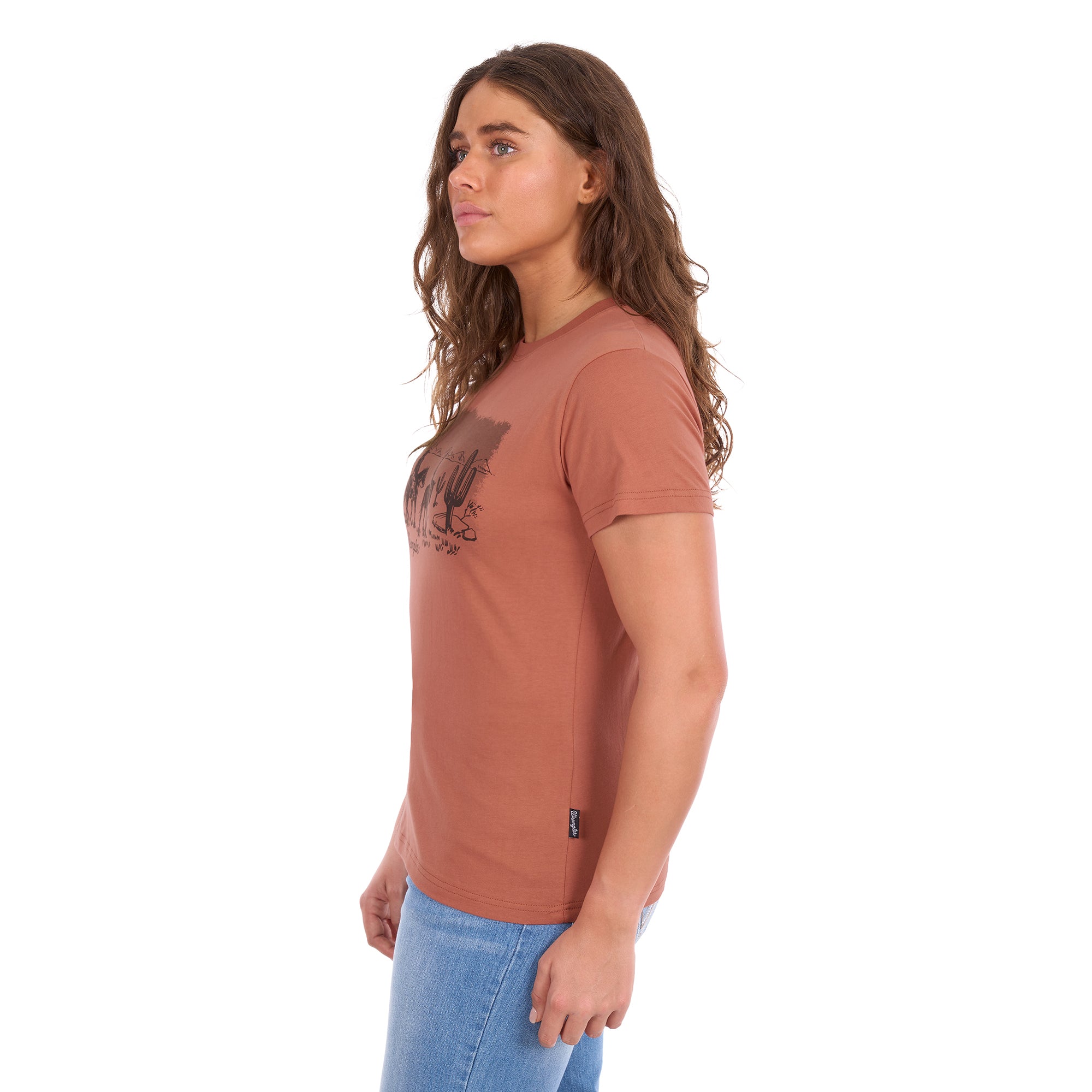 A woman wearing a copper coloured Wrangler Women's Adele S/S Tee (X6S2598644) with a dark graphic print on the front, paired with light blue jeans, shown in profile against a plain white background.