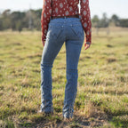 Rear view of a woman wearing faded blue Wrangler Women's Chara Willow Jeans (XCP2251216) with decorative stitching on the back pockets, a western shirt and brown cowboy boots out in a paddock with trees in the background.