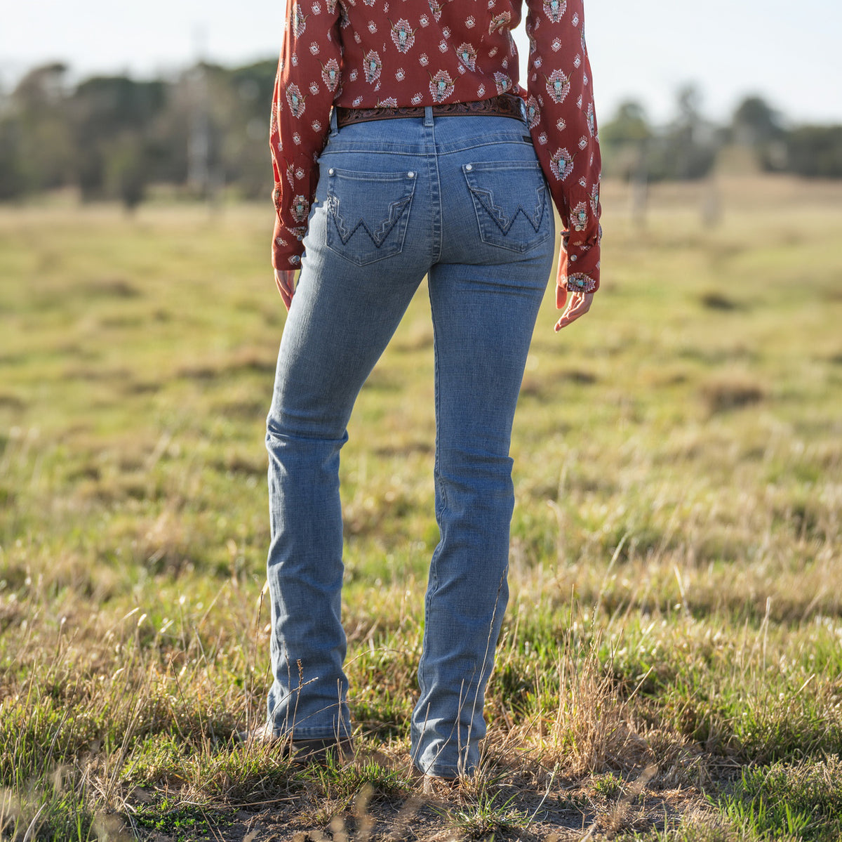 Rear view of a woman wearing faded blue Wrangler Women's Chara Willow Jeans (XCP2251216) with decorative stitching on the back pockets, a western shirt and brown cowboy boots out in a paddock with trees in the background.