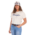 A woman modelling a Wrangler Women's Cindy Crop Short Sleeve T-Shirt (XCP2596647) in cream with a navy blue 'Wrangler' rope logo across the chest, paired with blue jeans, a Wrangler Nora High Profile Full Back Cap (XCP2947CAP) and a tan Wrangler Western Molly Belt (XCP2919BLT), against a plain white background.