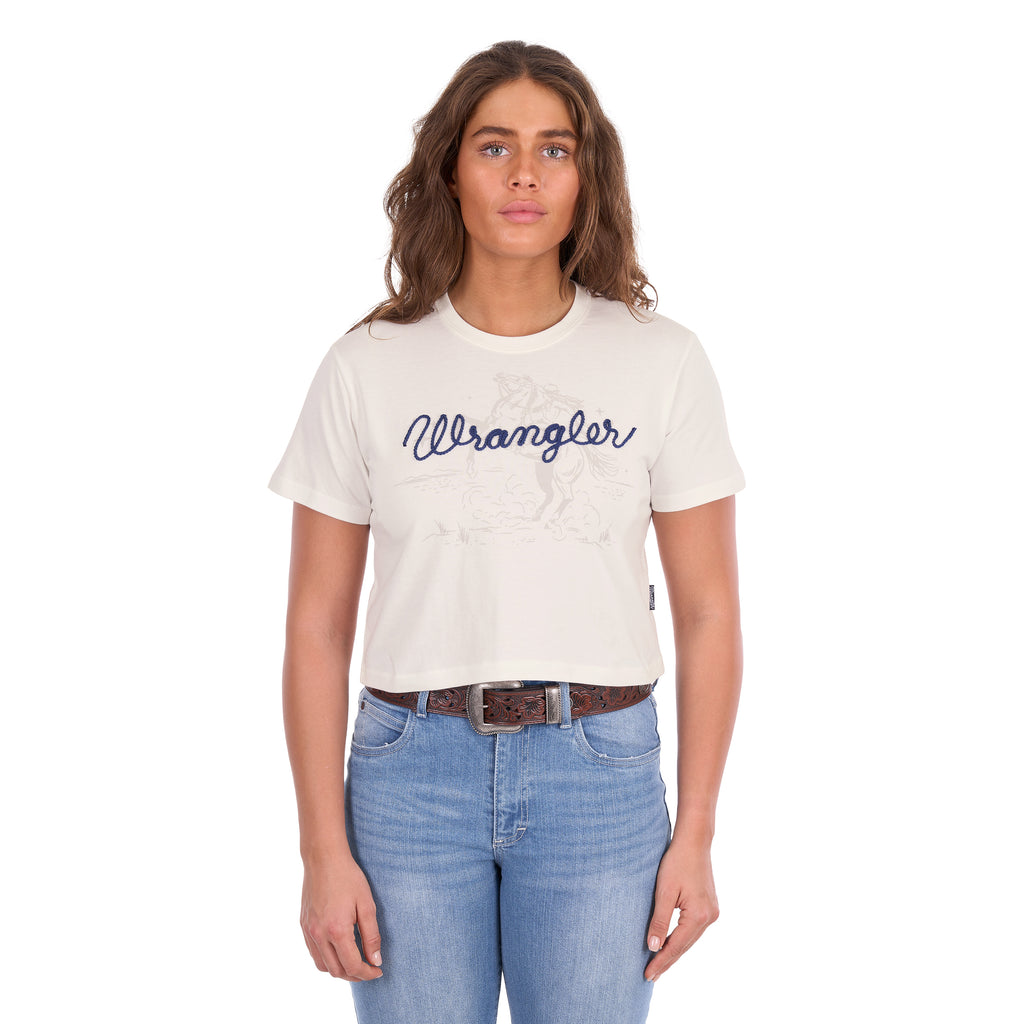 A woman modelling a Wrangler Women's Cindy Crop Short Sleeve T-Shirt (XCP2596647) in cream with a navy blue 'Wrangler' rope logo across the chest, paired with blue jeans and a tan Wrangler Western Molly Belt (XCP2919BLT), against a plain white background.