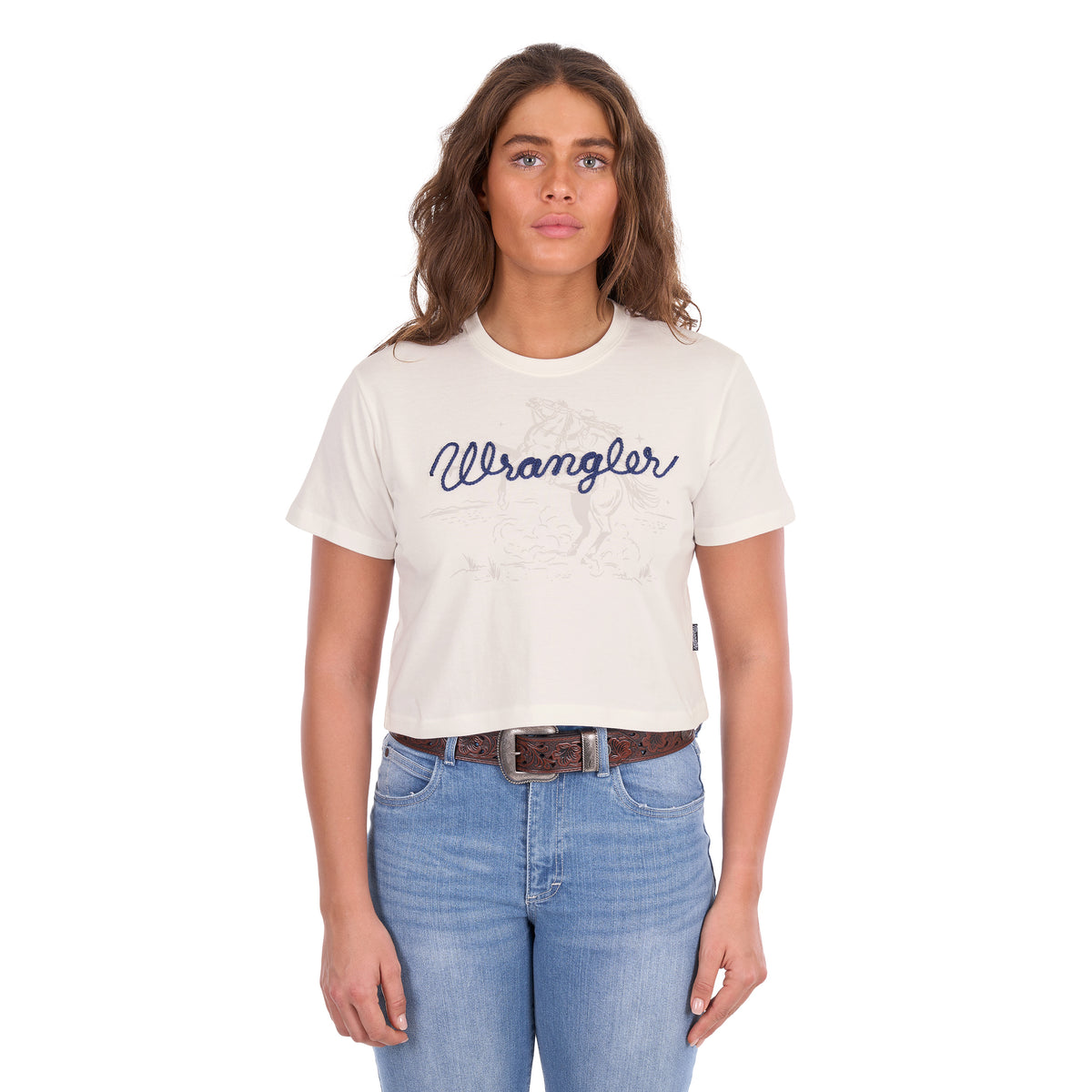 A woman modelling a Wrangler Women's Cindy Crop Short Sleeve T-Shirt (XCP2596647) in cream with a navy blue 'Wrangler' rope logo across the chest, paired with blue jeans and a tan Wrangler Western Molly Belt (XCP2919BLT), against a plain white background.