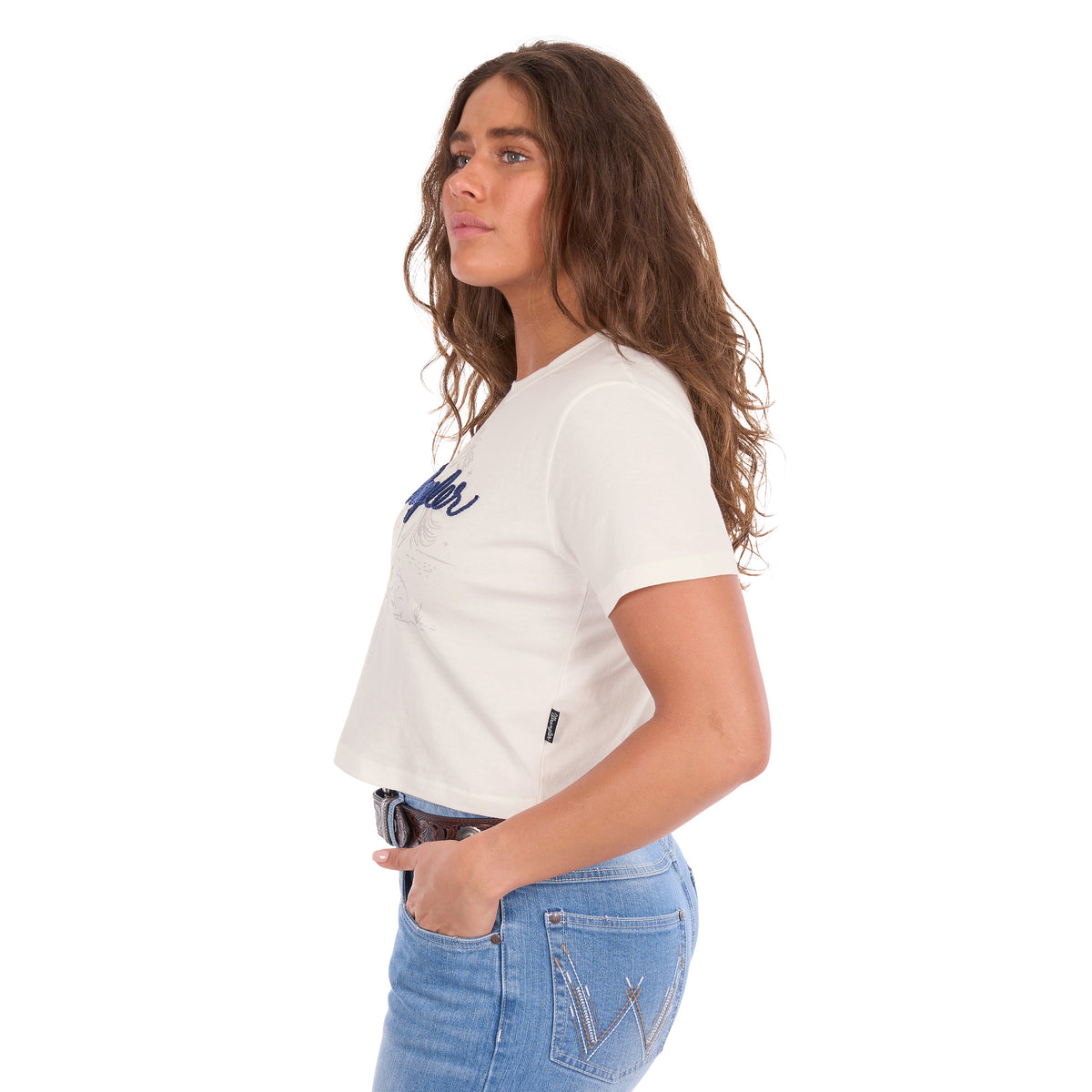 A woman in profile modelling a Wrangler Women's Cindy Crop Short Sleeve T-Shirt (XCP2596647) in cream with a navy blue 'Wrangler' rope logo across the chest, paired with blue jeans and a tan Wrangler Western Molly Belt (XCP2919BLT), against a plain white background.