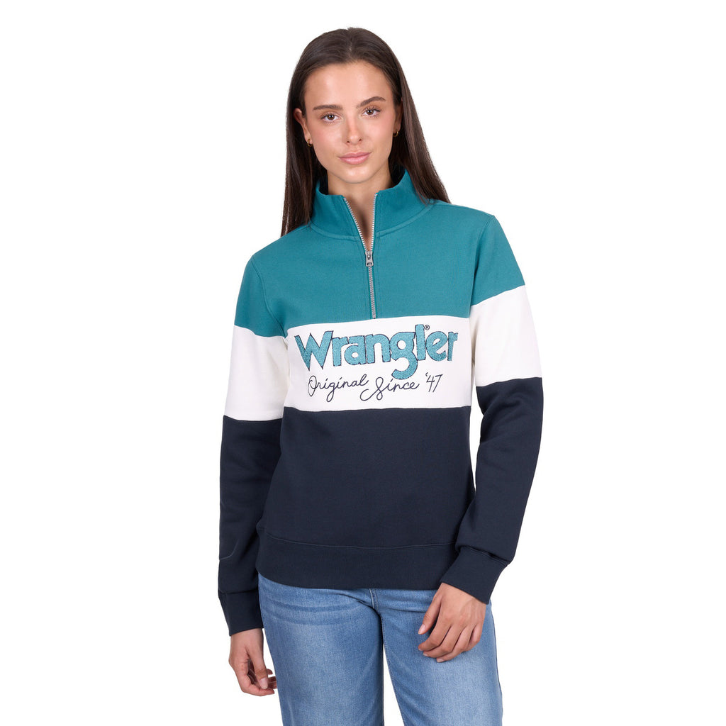 A woman stands against a white background wearing the Wrangler Womens Dana 1/4 Zip Pullover in Navy/Teal with a bold Wrangler logo, paired with light blue jeans.