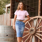 A Wrangler Women's Emmie Denim Skirt (X6S2410724) in light Kora Blue shown on a model, paired with a pastel pink Wrangler Women's Lucy Short Sleeved Tee (XCP2598633) and a tan Wrangler Siara Belt (X6S2916BLT) photographed outside a rustic shed with a wagon wheel and barrel.
