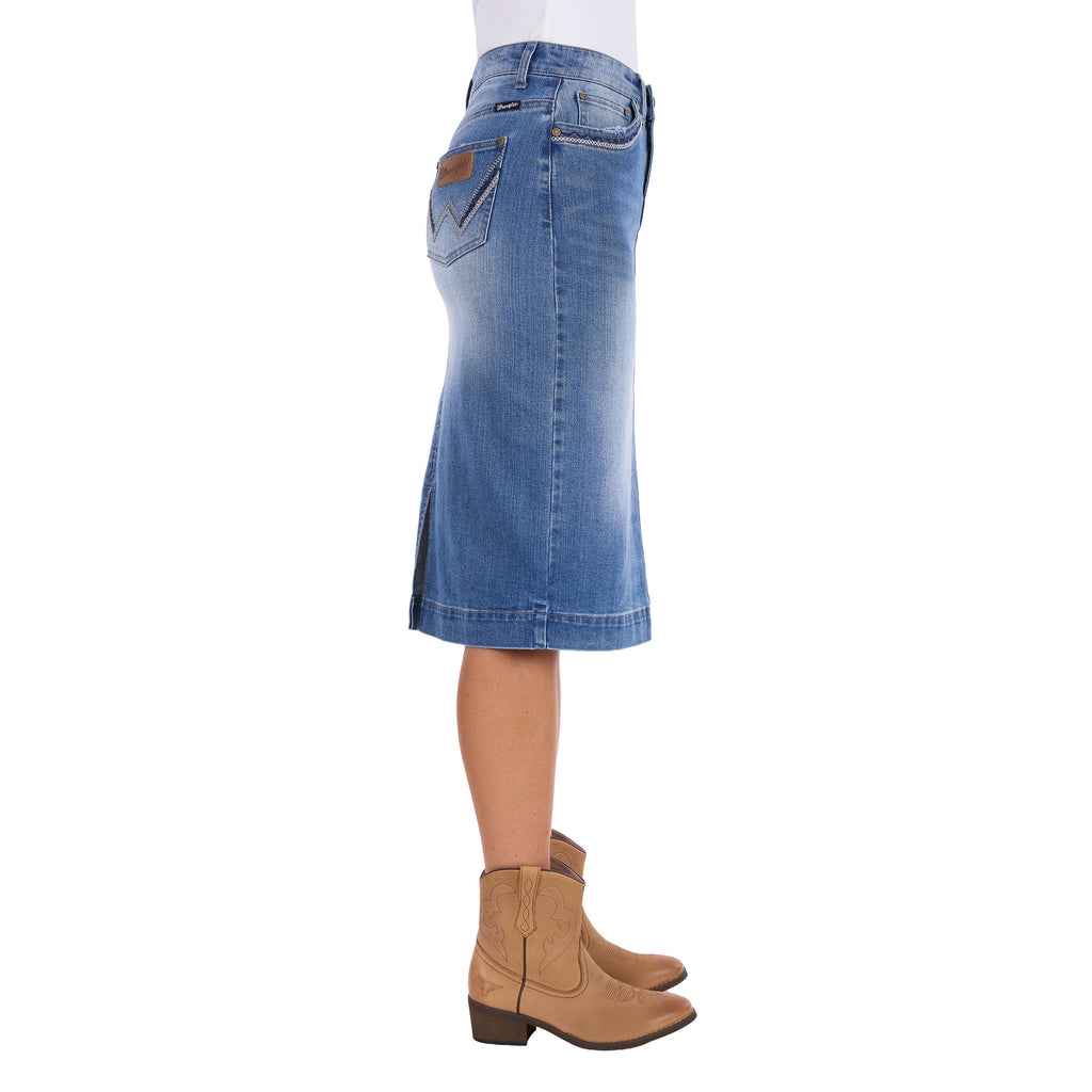 A Wrangler Women's Emmie Denim Skirt (X6S2410724) in light Kora Blue with embroidered pockets shown on a model from the waist down, photographed from the side, paired with tan cowboy boots against a plain white background.