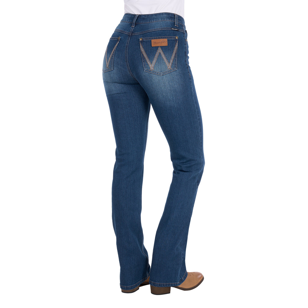 3-4 Rear view of a woman wearing mid indigo blue western-style Wrangler Women's Etta Bootcut Jeans Bailey (XCP2255711) with a white top and tan boots, shown against a plain white background.