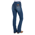 3-4 Rear view of a woman wearing mid indigo blue western-style Wrangler Women's Etta Bootcut Jeans Bailey (XCP2255711) with a white top and tan boots, shown against a plain white background.