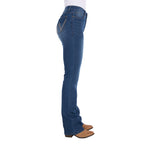 Side profile view of a woman wearing mid indigo blue western-style Wrangler Women's Etta Bootcut Jeans Bailey (XCP2255711) with a white top and tan boots, shown against a plain white background.