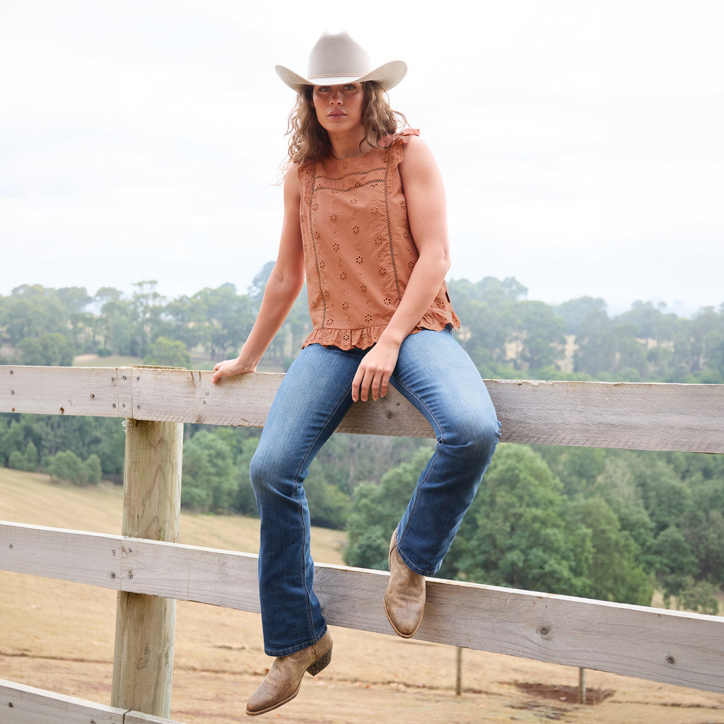 A woman wearing a copper Wrangler Women's Grace Sleeveless Blouse (X6S2502615) with eyelet embroidery and a ruffled hem, paired with blue jeans, and a silverton coloured Wrangler Brodie Cowboy Hat (XCP1961HAT) sits on a wooden fence in an outdoor rural setting.