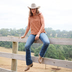 A woman wearing a copper Wrangler Women's Grace Sleeveless Blouse (X6S2502615) with eyelet embroidery and a ruffled hem, paired with blue jeans, and a silverton coloured Wrangler Brodie Cowboy Hat (XCP1961HAT) sits on a wooden fence in an outdoor rural setting.