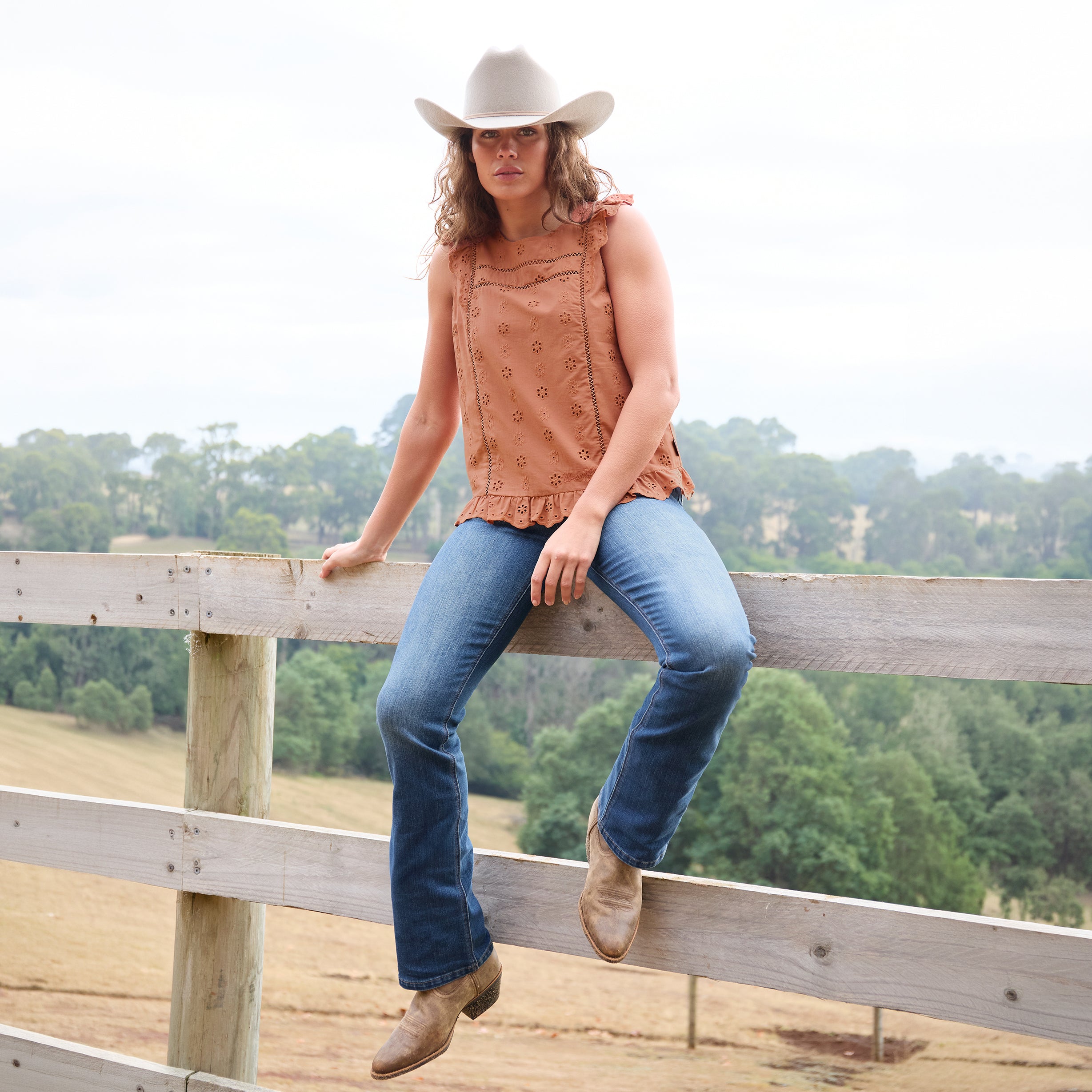 A woman wearing a copper Wrangler Women's Grace Sleeveless Blouse (X6S2502615) with eyelet embroidery and a ruffled hem, paired with blue jeans, and a silverton coloured Wrangler Brodie Cowboy Hat (XCP1961HAT) sits on a wooden fence in an outdoor rural setting.