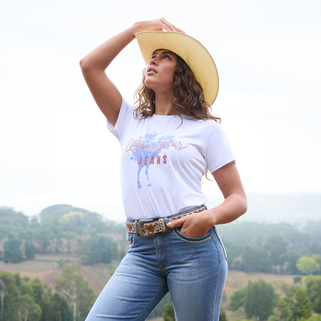 A woman wearing a white Wrangler Women's Gwen Tee (X6S2598635) with kora blue Wrangler Women's Margo Trouser Jeans Willow (XCP2253710), a Wrangler Blakely Belt (XCP2915BLT) and a Wrangler Jhonson Bangora (X4Y2025JHO) cowboy hat, posing outdoors against a soft-focus landscape background.