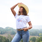 A woman wearing a white Wrangler Women's Gwen Tee (X6S2598635) with kora blue Wrangler Women's Margo Trouser Jeans Willow (XCP2253710), a Wrangler Blakely Belt (XCP2915BLT) and a Wrangler Jhonson Bangora (X4Y2025JHO) cowboy hat, posing outdoors against a soft-focus landscape background.