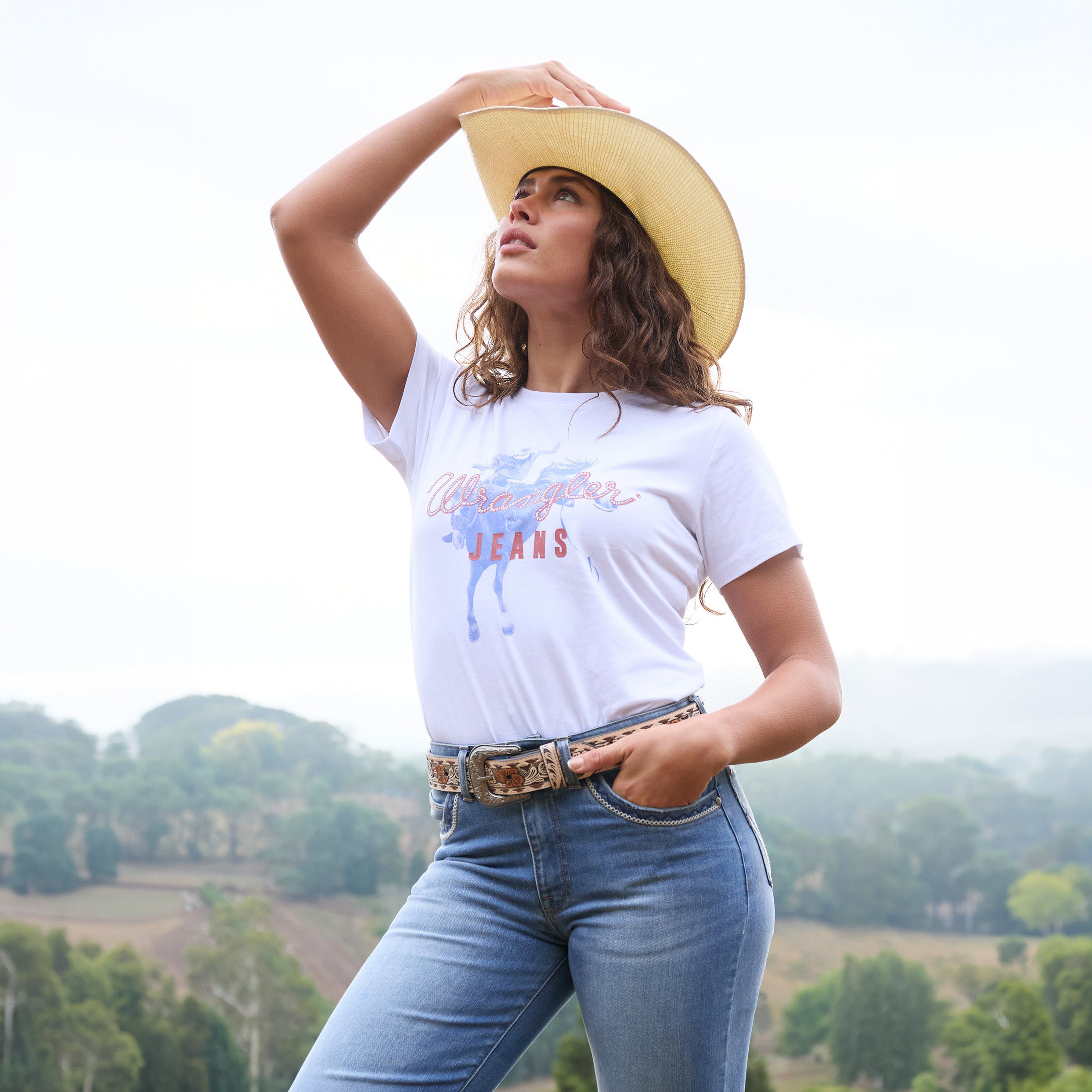 A woman wearing a white Wrangler Women's Gwen Tee (X6S2598635) with kora blue Wrangler Women's Margo Trouser Jeans Willow (XCP2253710), a Wrangler Blakely Belt (XCP2915BLT) and a Wrangler Jhonson Bangora (X4Y2025JHO) cowboy hat, posing outdoors against a soft-focus landscape background.