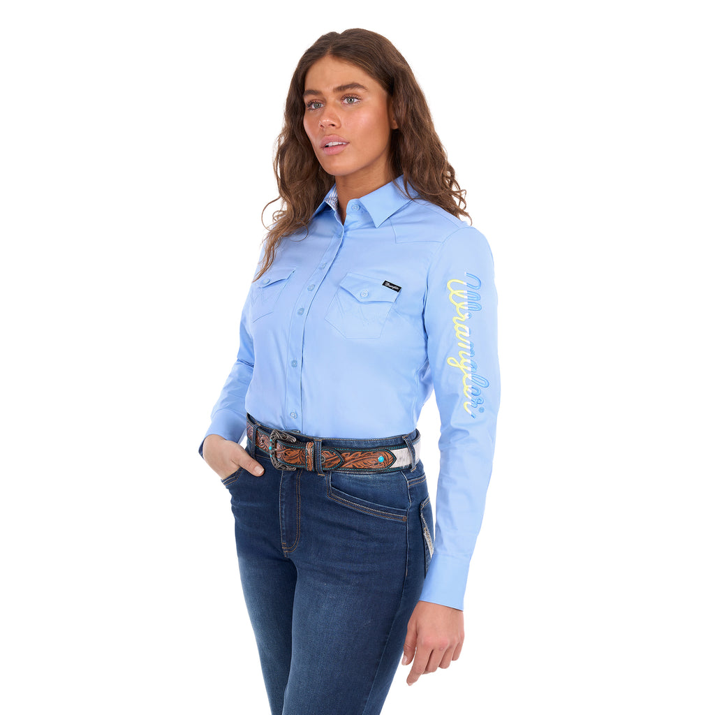 A woman modelling a blue Wrangler Women's Hazel Logo Long Sleeved Shirt (XCP2111608) with blue and cream Wrangler script embroidery on the right sleeve, paired with blue jeans and a brown western belt against a plain white background.