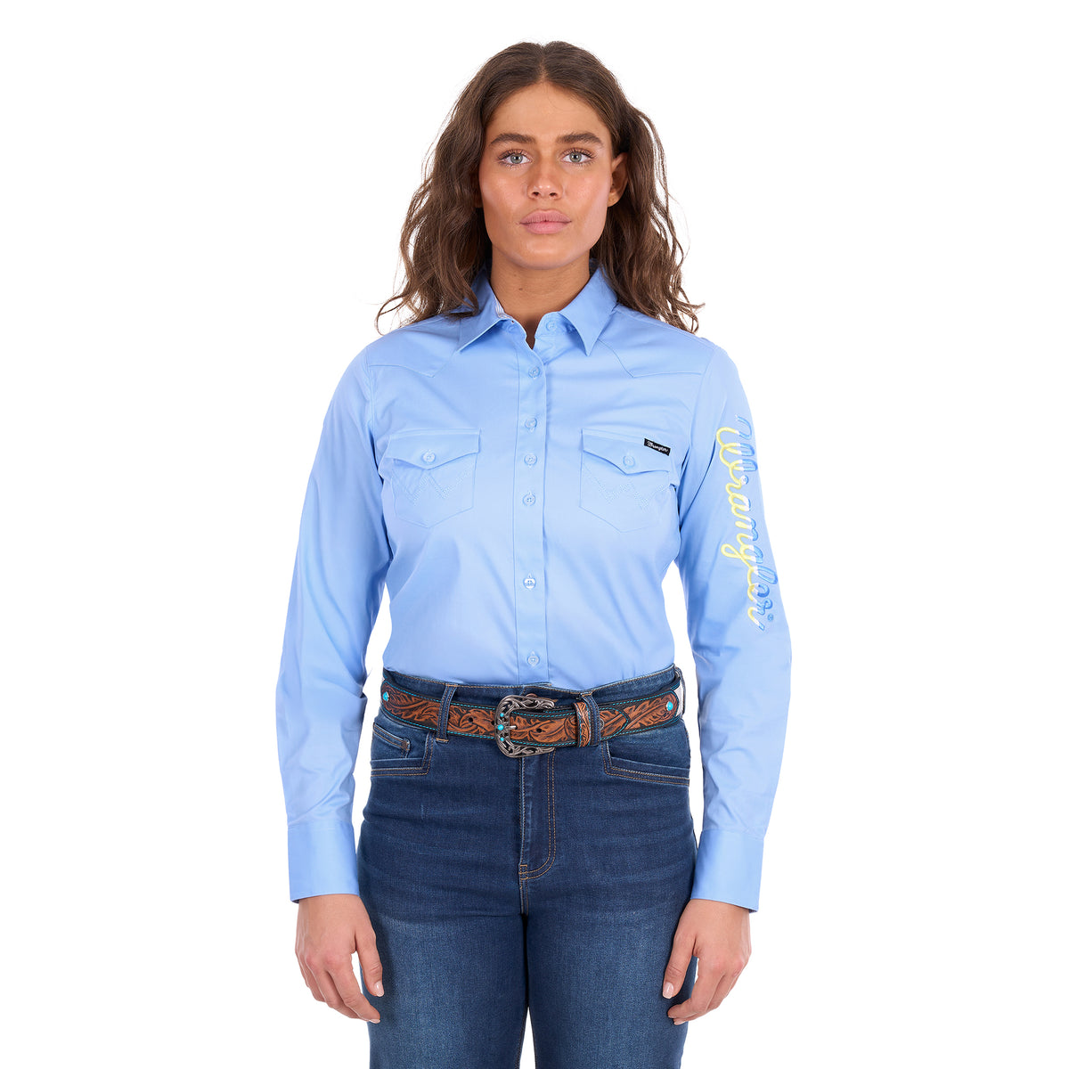 A woman modelling a blue Wrangler Women's Hazel Logo Long Sleeved Shirt (XCP2111608) with blue and cream Wrangler script embroidery on the right sleeve, paired with blue jeans and a brown western belt against a plain white background.