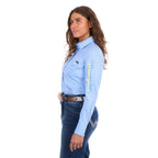 A woman modelling a blue Wrangler Women's Hazel Logo Long Sleeved Shirt (XCP2111608) with blue and cream Wrangler script embroidery on the right sleeve, paired with blue jeans and a brown western belt against a plain white background.