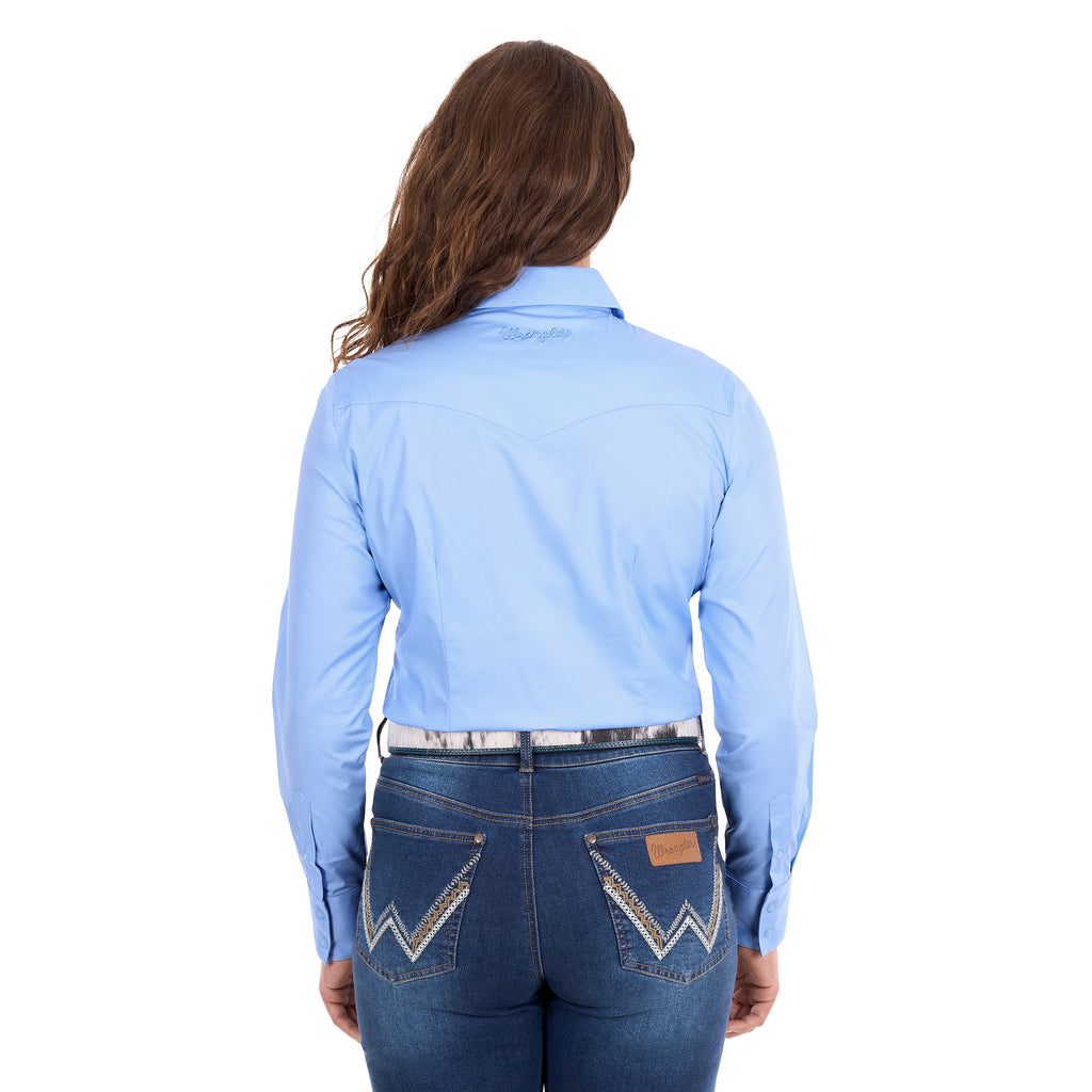 A woman modelling a blue Wrangler Women's Hazel Logo Long Sleeved Shirt (XCP2111608) with blue Wrangler script embroidery on the back neck, paired with blue jeans and a brown western belt against a plain white background.