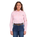 A woman modelling a blush pink Wrangler Women's Hazel Logo Long Sleeved Shirt (XCP2111608) with copper and pink Wrangler script embroidery on the right sleeve, paired with blue jeans and a brown western belt against a plain white background.