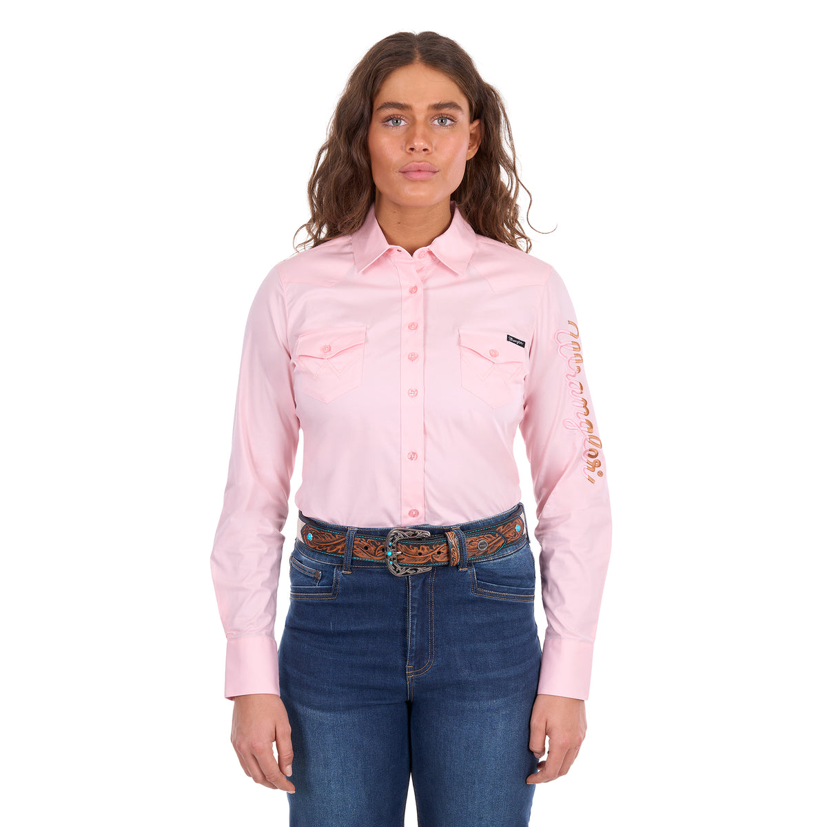 A woman modelling a blush pink Wrangler Women's Hazel Logo Long Sleeved Shirt (XCP2111608) with copper and pink Wrangler script embroidery on the right sleeve, paired with blue jeans and a brown western belt against a plain white background.