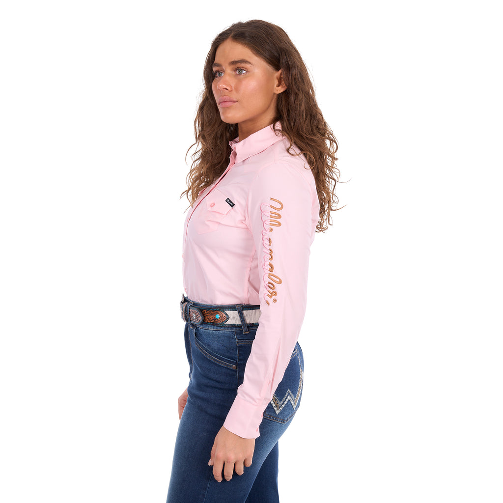 A woman modelling a blush pink Wrangler Women's Hazel Logo Long Sleeved Shirt (XCP2111608) with copper and pink Wrangler script embroidery on the right sleeve, paired with blue jeans and a brown western belt against a plain white background.
