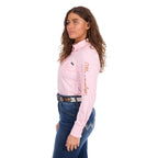 A woman modelling a blush pink Wrangler Women's Hazel Logo Long Sleeved Shirt (XCP2111608) with copper and pink Wrangler script embroidery on the right sleeve, paired with blue jeans and a brown western belt against a plain white background.