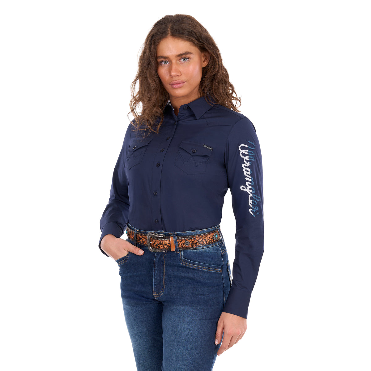 A woman modelling a navy Wrangler Women's Hazel Logo Long Sleeved Shirt (XCP2111608) with blue and white Wrangler script embroidery on the right sleeve, paired with blue jeans and a brown western belt against a plain white background.