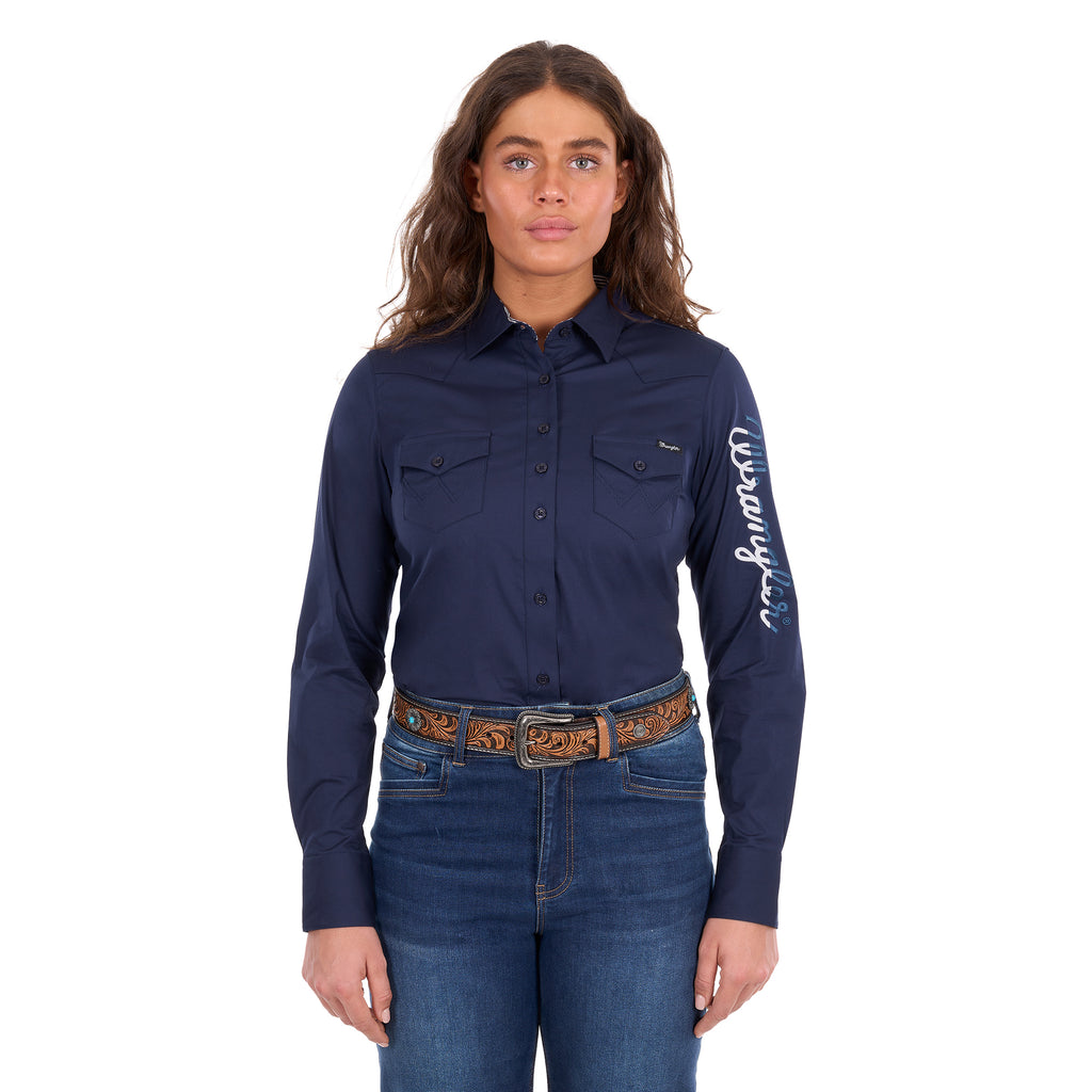 A woman modelling a navy Wrangler Women's Hazel Logo Long Sleeved Shirt (XCP2111608) with blue and white Wrangler script embroidery on the right sleeve, paired with blue jeans and a brown western belt against a plain white background.