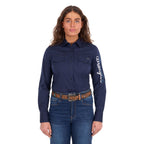 A woman modelling a navy Wrangler Women's Hazel Logo Long Sleeved Shirt (XCP2111608) with blue and white Wrangler script embroidery on the right sleeve, paired with blue jeans and a brown western belt against a plain white background.