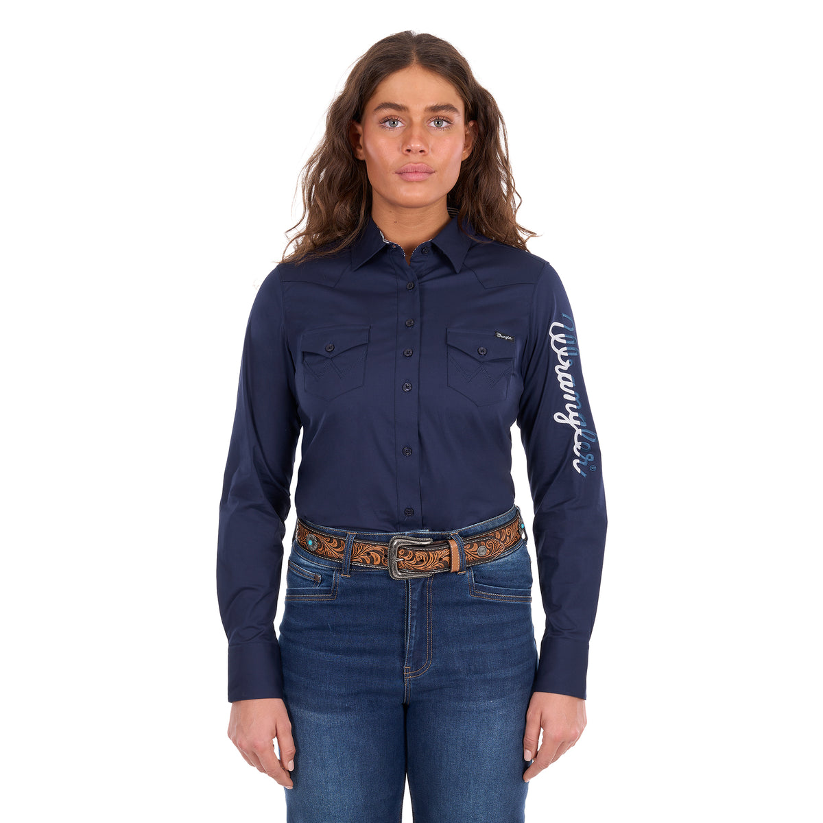 A woman modelling a navy Wrangler Women's Hazel Logo Long Sleeved Shirt (XCP2111608) with blue and white Wrangler script embroidery on the right sleeve, paired with blue jeans and a brown western belt against a plain white background.
