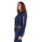 A woman modelling a navy Wrangler Women's Hazel Logo Long Sleeved Shirt (XCP2111608) with blue and white Wrangler script embroidery on the right sleeve, paired with blue jeans and a brown western belt against a plain white background.