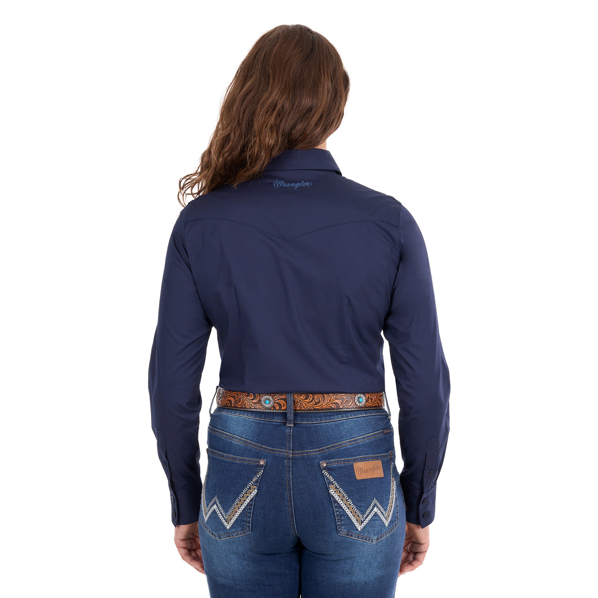 A woman modelling a navy Wrangler Women's Hazel Logo Long Sleeved Shirt (XCP2111608) with blue Wrangler script embroidery on the back neck, paired with blue jeans and a brown western belt against a plain white background.