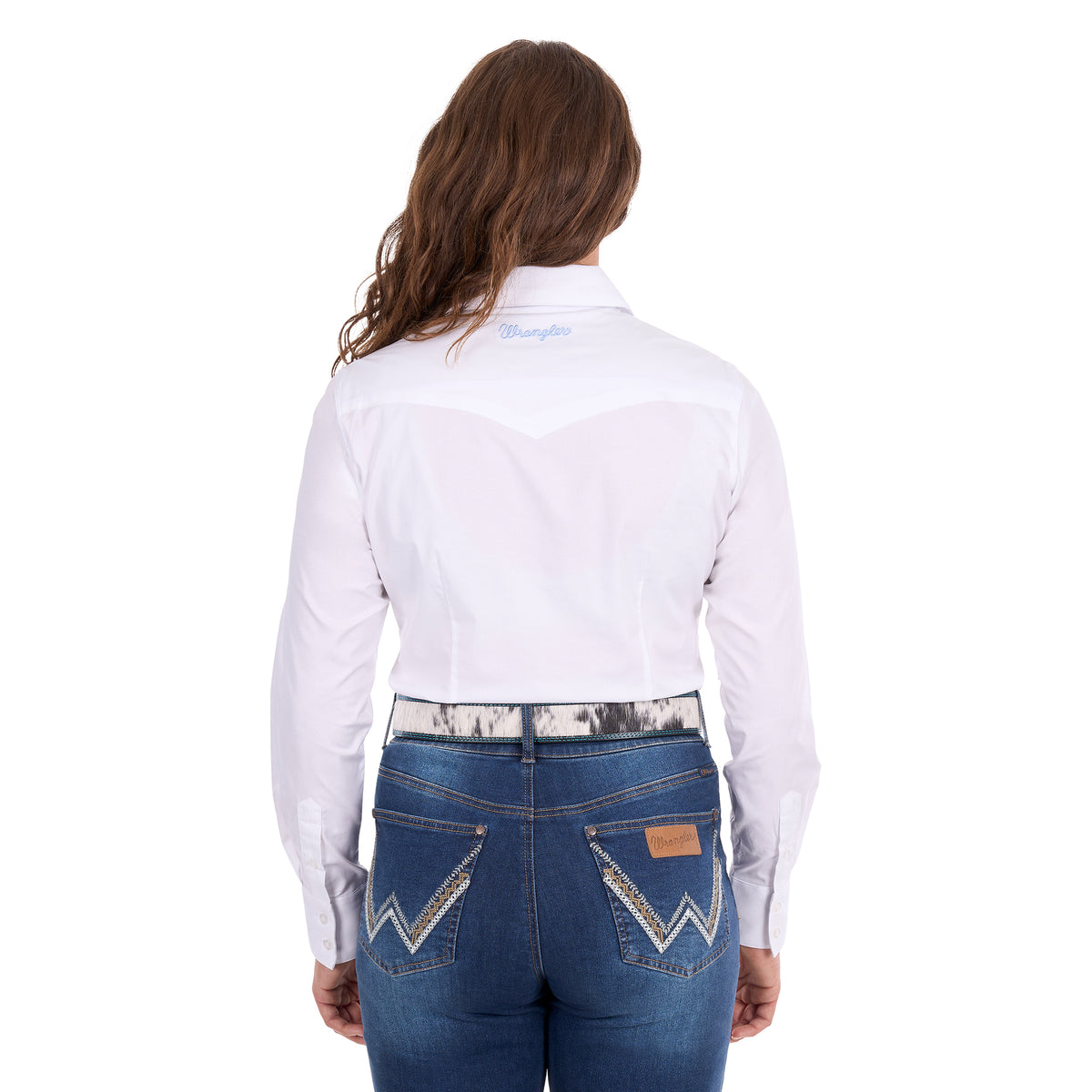 A woman modelling a white Wrangler Women's Hazel Logo Long Sleeved Shirt (XCP2111608) with blue Wrangler script embroidery on the back neck, paired with blue jeans and a western belt against a plain white background.
