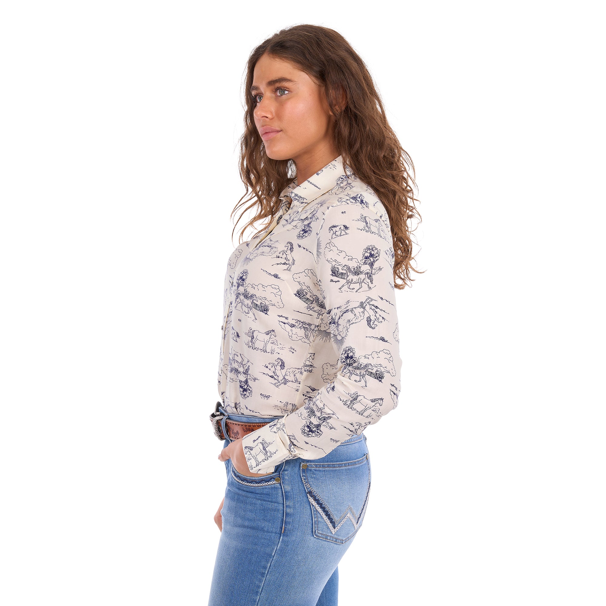 A woman in profile modelling a cream Wrangler Women's Ivory Long Sleeve Shirt (X6S2103602) with a western novelty print paired with kora blue Wrangler Women's Margo Trouser Jeans Willow (XCP2253710) and a Wrangler Silvie Belt (X6S2920BLT), against a plain white studio background.