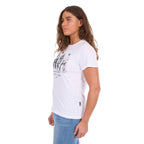 A woman in profile modelling a white graphic Wrangler Women's Josie Short Sleeve T-Shirt (XCP2598641) with a black and white print of riders on a fence and the text 'Wrangler Butts Drive Me Nuts' on the front, paired with blue jeans against a plain white background.