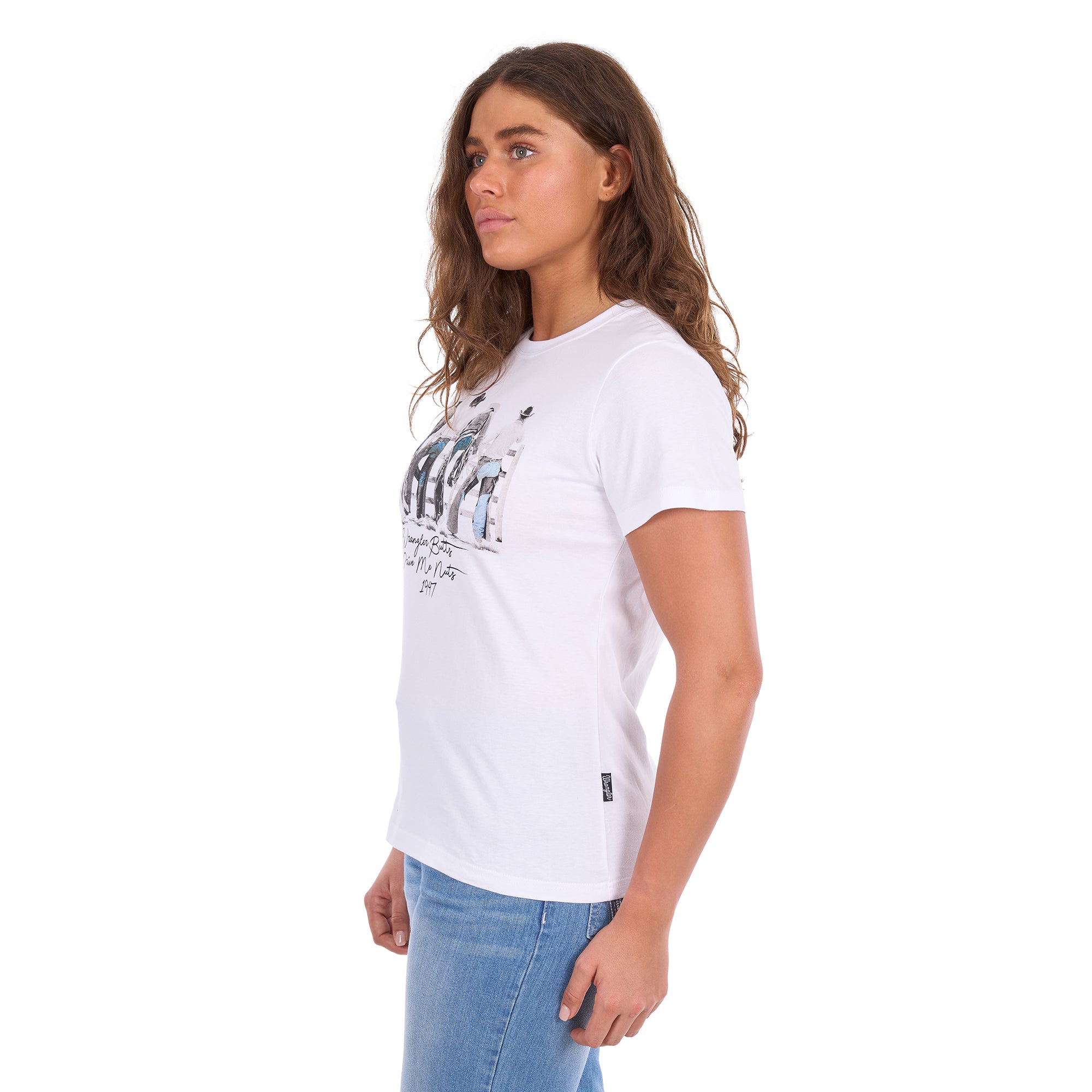 A woman in profile modelling a white graphic Wrangler Women's Josie Short Sleeve T-Shirt (XCP2598641) with a black and white print of riders on a fence and the text 'Wrangler Butts Drive Me Nuts' on the front, paired with blue jeans against a plain white background.