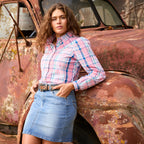 A woman wearing a pink, blue, and white plaid Wrangler Kiara Long Sleeve Shirt (X6S2105603) a tan Wrangler Siara Belt (X6S2916BLT) and a light blue Wrangler Maisy Denim Skirt (XCP2409723), leaning against a rusted vintage car.