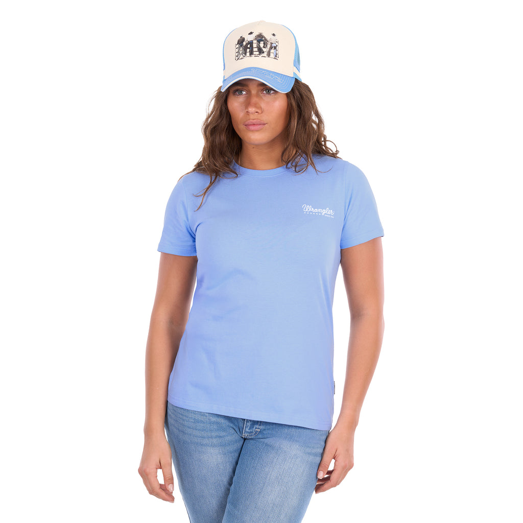 A woman facing the camera wearing a light blue crewneck Wrangler Women's Lola Short Sleeved Tee (X6S2598642) with a small white logo on the left chest, paired with a Wrangler Josie High Profile Trucker Cap (X6S2943CAP) in Cream and Light Blue, with blue jeans, against a solid white background.