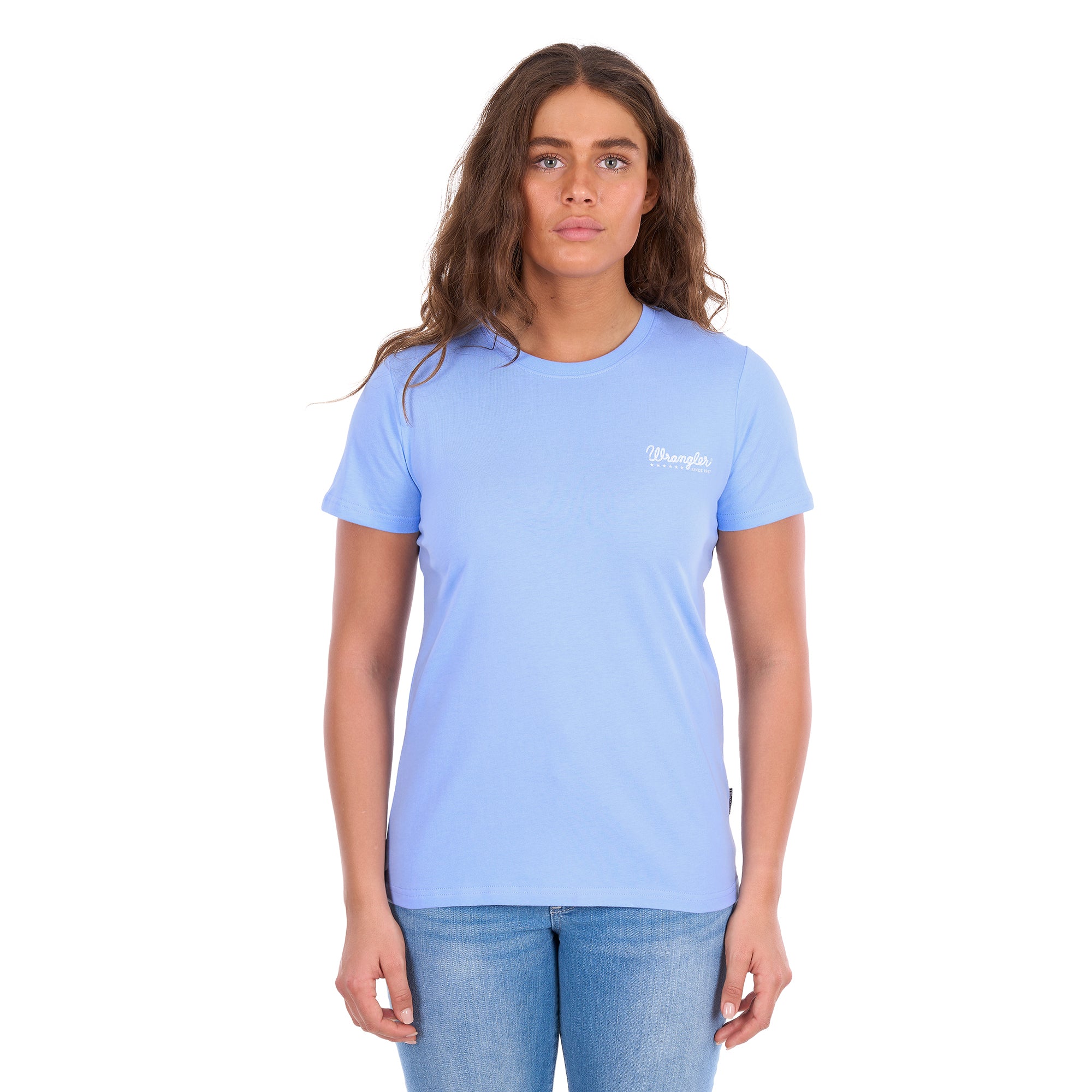 A woman facing the camera wearing a light blue crewneck Wrangler Women's Lola Short Sleeved Tee (X6S2598642) with a small white logo on the left chest, paired with blue jeans, against a solid white background.
