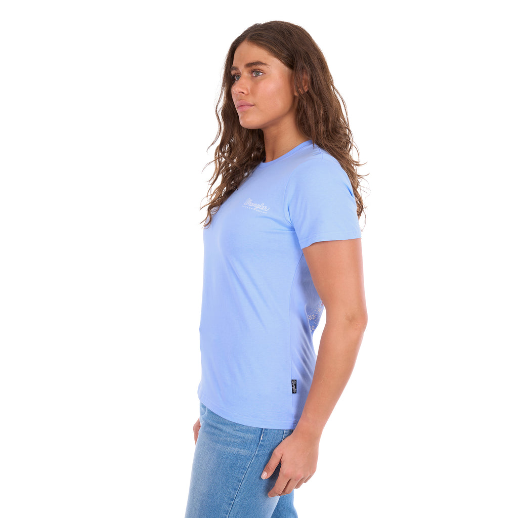 A woman wearing a light blue crewneck Wrangler Women's Lola Short Sleeved Tee (X6S2598642) with a small white logo on the left chest, paired with blue jeans, shown in side profile against a solid white background.